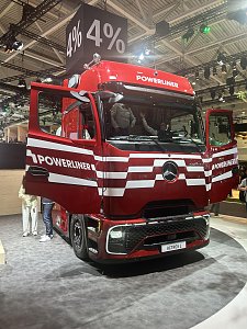 Dunkelroter Mercedes-Benz Lkw von vorne links auf einer Messe. Die Türen stehen offen. Auf der Front sind weiße Querstreifen und der Schriftzug POWERLINER zu sehen. Über dem Lkw hängt ein schwarzer Würfel mit der weißen Aufschrift 4%.