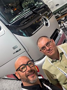 Selfie von zwei Männern mit Brille auf einer Messe. Im Hintergrund steht die silberne Front eines Mercedes-Benz Lkws mit großem Logo. Der Mann vorne lächelt glücklich, der Mann dahinter schaut ruhig in die Kamera.