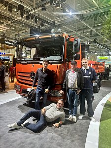 Vier Männer posieren glücklich vor einem großen, orangefarbenen Mercedes-Benz Lkw auf einer Messe. Drei Männer stehen aufrecht neben der Stoßstange, während der vierte Mann entspannt auf dem grauen Teppichboden davor liegt.