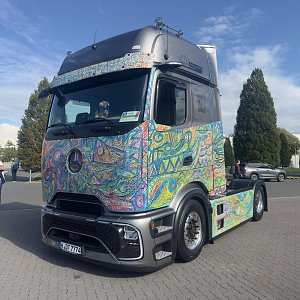 Bunt bemalter Mercedes-Benz Lkw von vorne links auf einem Pflasterplatz. Die Karosserie ist mit wilden, farbenfrohen Mustern bedeckt. Das Dach und die Stoßstange sind mattgrau. Im Hintergrund blauer Himmel.
