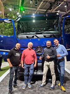 Vier lächelnde Männer posieren eng nebeneinander vor einem großen, blauen Mercedes-Benz Lkw auf einer Messe. Die beiden Türen des Lastwagens stehen weit offen.