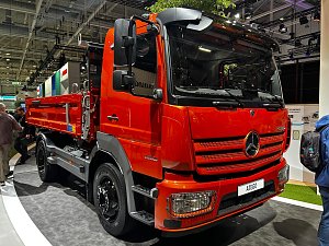 Roter Mercedes-Benz Kipper-Lkw von vorne rechts auf einem Messestand. Das Fahrzeug hat ein kurzes Fahrerhaus, schwarze Felgen und eine rote Ladefläche hinten. Personen laufen im Hintergrund vorbei. Der Lkw steht auf grauem Teppich.