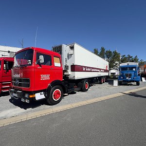Roter historischer Mercedes-Benz Sattelzug von vorne links auf einem asphaltierten Platz. Der weiße Auflieger hat einen braunen Streifen. Rote Felgen. Im Hintergrund rechts steht ein blauer Oldtimer-Lkw unter wolkenlosem, blauem Himmel.