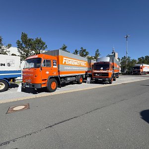 Orangefarbener Oldtimer-Lkw von vorne links auf einem Parkplatz. Er hat eine graue Plane mit der Aufschrift FERNVERKEHR. Dahinter stehen zwei weitere, baugleiche Lastwagen in orange.
