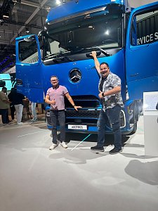 Zwei lachende Männer posieren vor einem großen, blauen Mercedes-Benz Lkw auf einer Messe. Beide Türen des Lastwagens stehen weit offen.