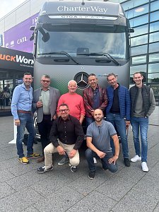Acht Männer posieren lächelnd vor einem großen, grauen Mercedes-Benz Lkw auf einem Außenbereich. Sechs Männer stehen aufrecht nebeneinander, während zwei Männer davor in der Hocke sitzen. Oben auf dem Lkw steht der weiße Schriftzug CharterWay.