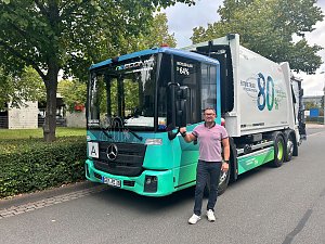 Ein Mann steht lächelnd neben einem modernen, hellgrün-blauen Mercedes-Benz Müllwagen. Der weiße Lkw-Aufbau hinten trägt die große Aufschrift 80%. Das Fahrzeug steht am Straßenrand vor grünen Bäumen und einer Hecke.