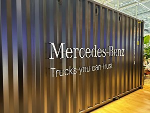 Dunkelgraue Containerwand aus Wellblech auf einer Messe. In der Mitte prallt der silberne, erhabene Schriftzug Mercedes-Benz hervor. Darunter steht der weiße Slogan Trucks you can trust.
