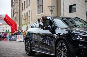 Man sieht das Führungsfahrzeug, den vollelektrischen GLC. Aus dem Beifahrerfenster heraus fotografiert eine Frau. Das Auto steht. Im Hintergrund sind die Läufer und Läuferinnen zu sehen.