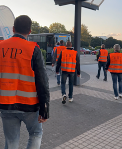 Mehrere Personen von hinten gesehen gehen in leuchtend orangefarbenen VIP-Warnwesten über ein Außengelände auf einen bereitstehenden Bus zu.