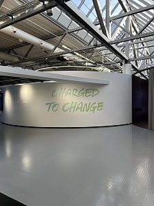 Eine geschwungene, weiße Wand steht in einer modernen Halle mit glänzendem, grauem Boden und einer offenen, verglasten Dachkonstruktion aus Stahlträgern. Auf der Wand prangt der hellgrüne Schriftzug CHARGED TO CHANGE.