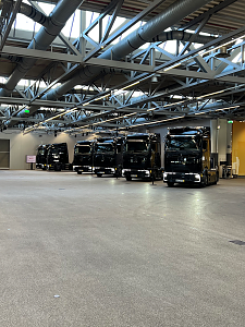 Eine ganze Reihe fabrikneuer, glänzend schwarzer Mercedes-Benz Lkw steht nebeneinander aufgereiht in einer riesigen Werkshalle.