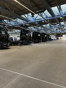 Mehrere schwarze Mercedes-Benz Lkw-Zugmaschinen stehen schräg hintereinander aufgereiht in einer weiten Produktionshalle. Der Blickwinkel zeigt die Fahrzeuge von der Seite auf einer grauen Betonfläche.