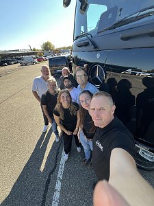 Eine Gruppe von acht Personen posiert für ein Selfie direkt neben der glänzenden, schwarzen Front eines Mercedes-Benz Lkw.