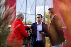 Drei Männer im Gespräch bei der Fahrzeugübergabe an den Thüringer HC, darunter ein Mann im dunkelblauen Anzug mit Smartphone.