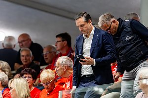 Ein Mann im dunkelblauen Anzug blickt auf sein Smartphone, während Zuschauer in roten Trikots auf der Tribüne der Handballhalle sitzen.