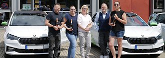 Fünf Personen stehen vor einem modernen Gebäude und posieren mit Champagnerflaschen vor zwei weißen Škoda-Fahrzeugen. Auf den Autos sind grüne Elemente und das Logo der Praxisklinik für Zahnheilkunde zu sehen. (Autohaus Peter)