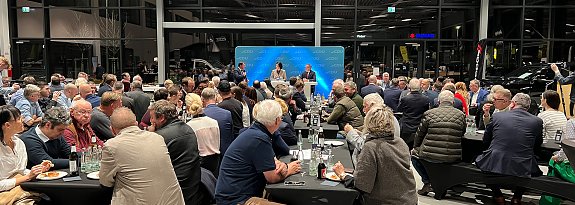 Eine große Gruppe von Menschen sitzt an langen, schwarz gedeckten Tischen in einer modernen Autohaus-Halle. Vorne steht eine Bühne mit CDU-Logo, auf der zwei Personen sprechen. Im Hintergrund sind Glasfassaden und Autos sichtbar. (Autohaus Peter Gruppe)