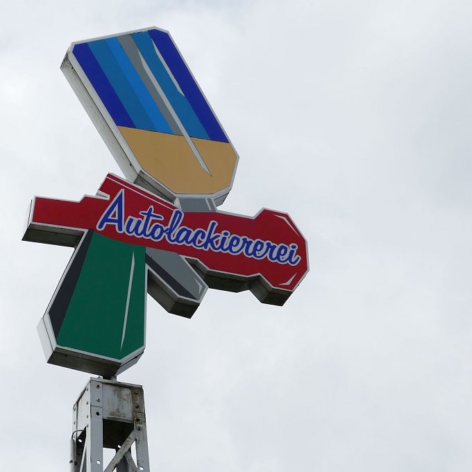 Werbeschild mit bunter Grafik in Form einer stilisierten Sprühflasche und dem Schriftzug Autolackiererei in blauer und roter Schrift. Der Himmel ist grau und bewölkt.
