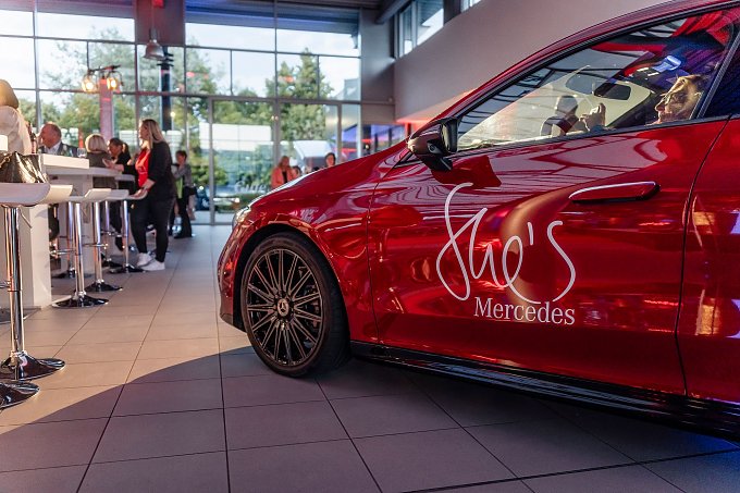 Ein rotes Mercedes-Fahrzeug mit der Aufschrift „Shes Mercedes“ auf der Fahrertür steht in einem hellen Showroom. Im Hintergrund unterhalten sich Menschen an Stehtischen. Mercedes-Benz CLA 250+ mit EQ-Technologie | Energieverbrauch kombiniert: 14,1-12,2 kWh/100 km | CO2Emissionen kombiniert: 0 g/km | CO2-Emissionsklasse: A