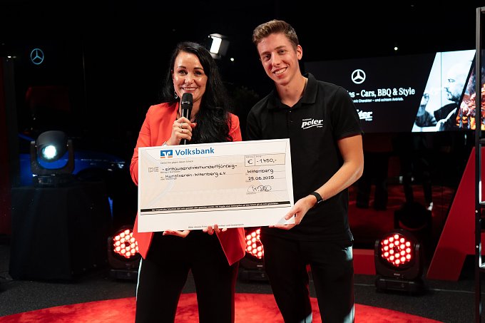 Christina Dost im roten Blazer und ein junger Mann im schwarzen Polohemd halten gemeinsam einen übergroßen Spendenscheck mit dem Logo der Volksbank.