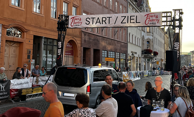 Zu sehen ist die Start Ziel Linie des Radsportrennen in Wittenberg. Darunter befindet sich ein silberner EQV, der als Führungsfahrzeug diente. Rund um die Start Ziel Linie sind hunderte Zuschauer zu sehen.
