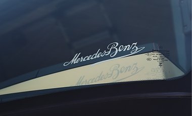 Nahaufnahme einer Windschutzscheibe. Im Glas ist der geschwungene, wei&szlig;e Schriftzug Mercedes-Benz in Schreibschrift eingelassen. Darunter wirft er einen Schatten auf das Armaturenbrett. Rechts stehen kleine Pr&uuml;fzeichen auf dem Glas.